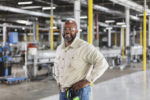 A SMILING MAN IN SAFETY GOGGLES STANDS, HANDS ON HIPS, IN FRONT OF A PRODUCTION LINE