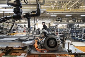 A shell of a car on a manufacturing floor, with a man in a black shirt standing behind it