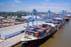 A black container ship docked at a port, under two large blue shipping cranes.