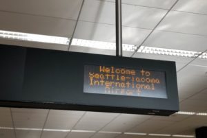 A black digital sign with yellow letters hanging from a ceiling, that reads "Welcome to Seattle-Tacoma International Airport"