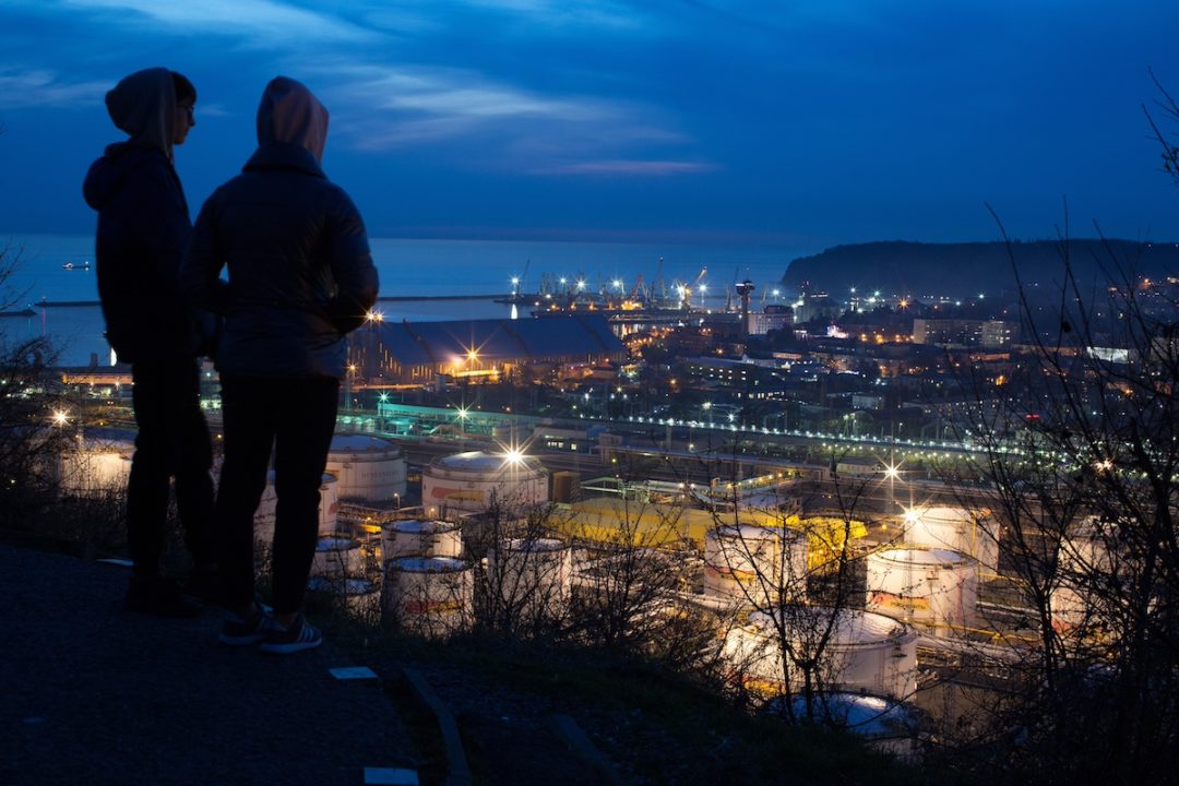 Two people silhouetted in the foreground, looking over a large industrial area at twilight.