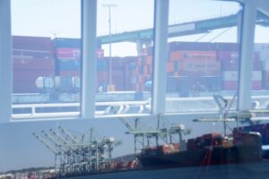 A window looking out at a container ship docked at a shoreline next to blue shipping cranes and stacks of containers