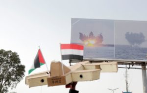 A cardboard model of an airplane with two small flags planted in it, with a billboard depicting explosions at sea in the background.