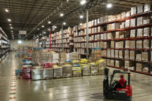 A WORKER DRIVES A FORKLIFT TRUCK IN THE FOREGROUND OF A VAST WAREHOUSE STACKED WITH PALLETS OF GOODS