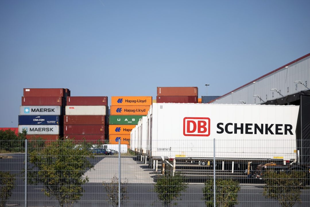 A row of white shipping containers with "DB Schenker" written on the outside, with a stack of multi-colored shipping containers in the background