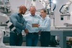 SEVERAL WORKERS CLUSTER AROUND A COMPUTER TABLET IN A ROOM FULL OF MANUFACTURING ROBOTS