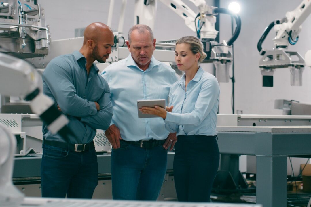 SEVERAL WORKERS CLUSTER AROUND A COMPUTER TABLET IN A ROOM FULL OF MANUFACTURING ROBOTS