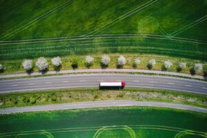 An overhead view of a white and red semi-truck driving on a rode alongside green grass fields