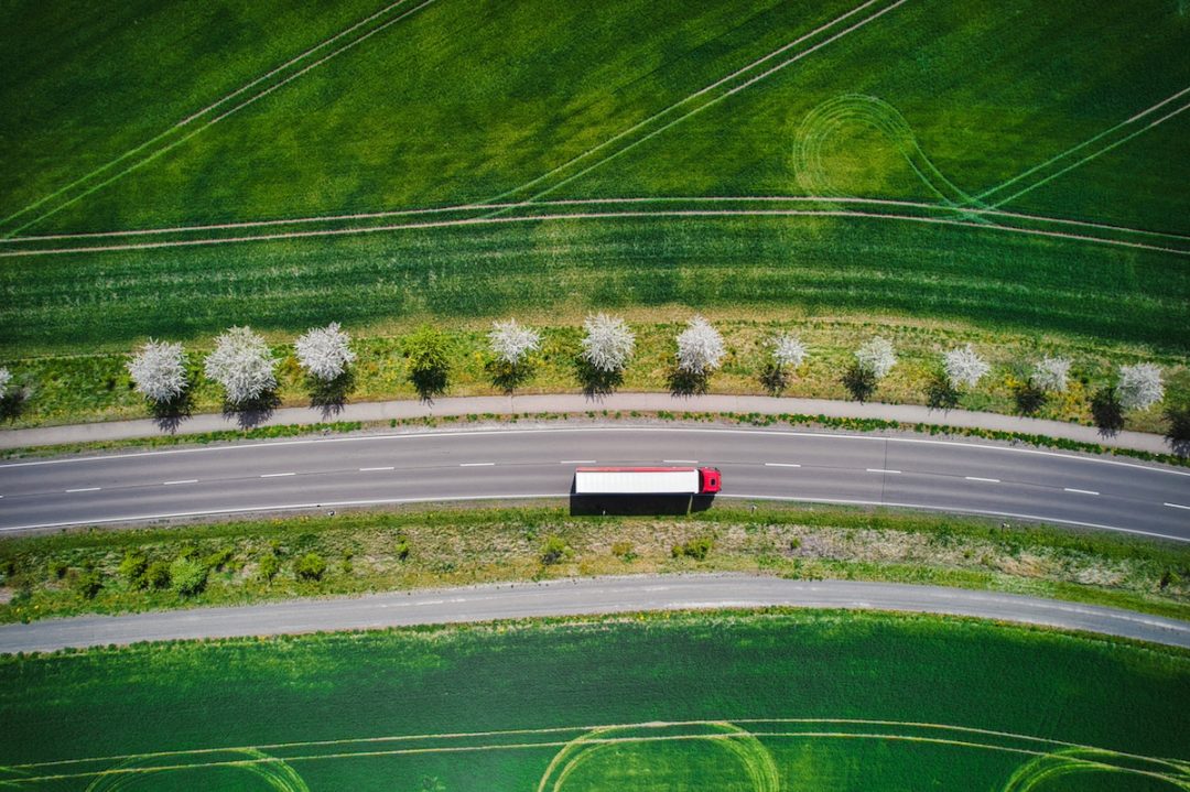 An overhead view of a white and red semi-truck driving on a rode alongside green grass fields