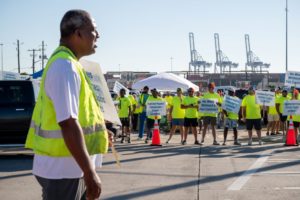 STRIKING WORKERS IN HI-VIS VESTS STAND OUTSIDE A PORT ENTRANCE