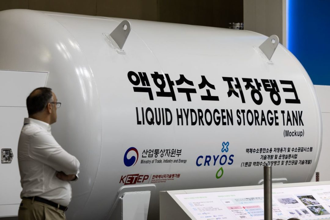 A MAN IN A WHITE SHIRT STANDS WITH FOLDED ARMS REGARDING A HYDROGEN STORAGE TANK PRINTED WITH BOTH ENGLISH AND KOREAN LETTERING.