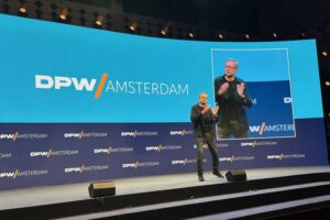  A MAN IN CASUAL BLACK CLOTHES ADDRESSES AN AUDIENCE FROM A STAGE THAT BEARS A DPW AMSTERDAM BACKDROP