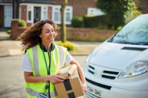 A YOUNG, SMILING WOMAN HOLDS A PACKAGE AS SHE WALKS FROM A DELIVERY VAN IN A SUBURBAN STREET06726727.jpg