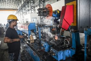 A man in a black t-shirt and a yellow hard hat standing in front of a large blue industrial machine on a factory assembly line.