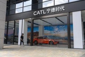 The front of a building with floor-to-ceiling glass windows and "CATL" written in white lettering on black trim, with an orange sports car directly inside the front glass doors