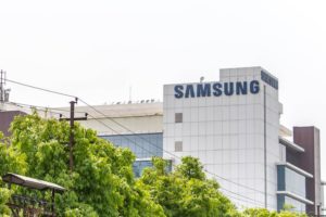 A white office building with "SAMSUNG" written in large blue letters at the top right, with a row of trees in the foreground