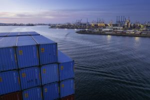 A LARGE PORT WITH LOTS OF CRANES IS VISIBLE OVER A STACK OF CONTAINERS IN THE DYING VIOLET LIGHT