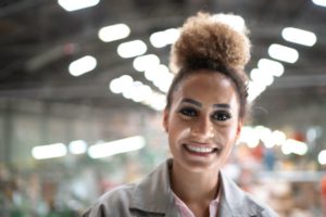 A SMILING WOMAN OF COLOR STANDS IN A BLURRY WAREHOUSE ENVIRONMENT