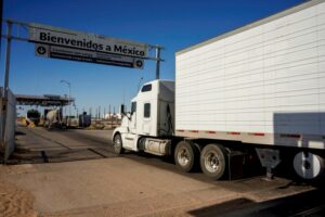 A TRUCK ON A RED TARMAC ROAD APPROACHES AN OVERHEAD SIGN THAT READS BIENVENIDOS A MEXICO