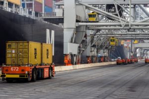 An orange, driverless vehicle moving two yellow shipping containers down a road, with a large black container ship docked alongside the road on the left