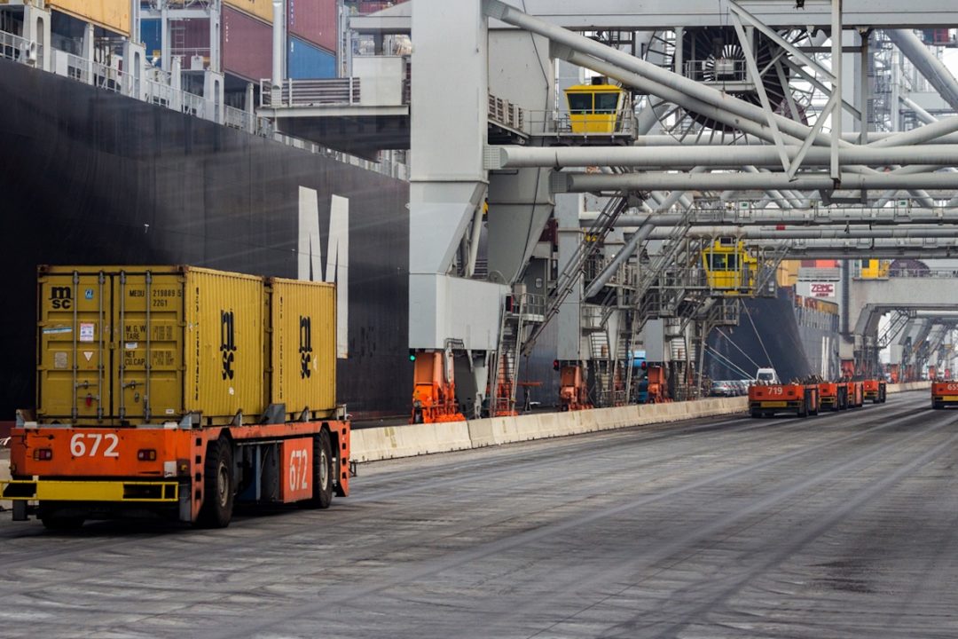 An orange, driverless vehicle moving two yellow shipping containers down a road, with a large black container ship docked alongside the road on the left