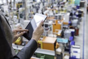 A view of a woman's hands holding an tablet, with orange and blue factory machines blurred out in the background below