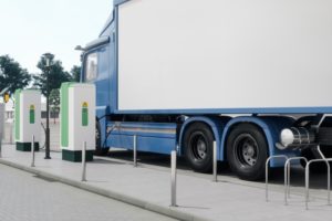 A blue semi-truck with a white cab, parked along a curb next to two white and green charging boxes.