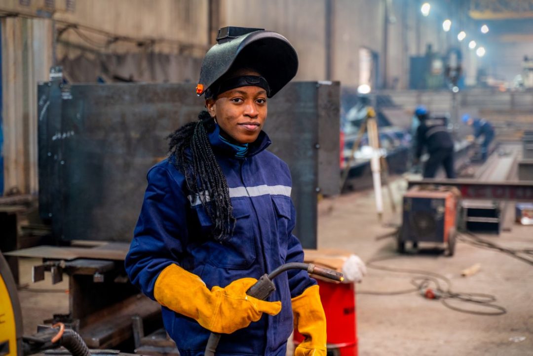 A WOMAN OF COLOR IN BLUE OVERALLS HOLDS A WELDING TORCH IN A FACTORY SETTING