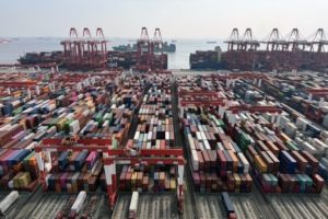 Dozens of rows of multi-colored shipping containers stacked on top of each other, with a row of red shipping cranes in the background