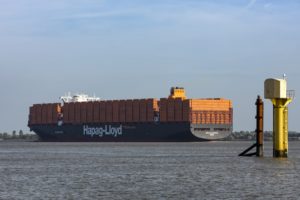 A black container ships with "Hapag-Lloyd" written along the side in large white letters, carrying dozens of stacks of orange shipping containers. 