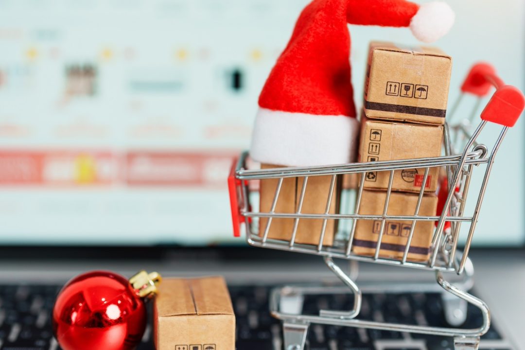 A miniature shopping car on a laptop keyboard, with four brown cardboard boxes and a red Santa hat, next to a round red Christmas ornament