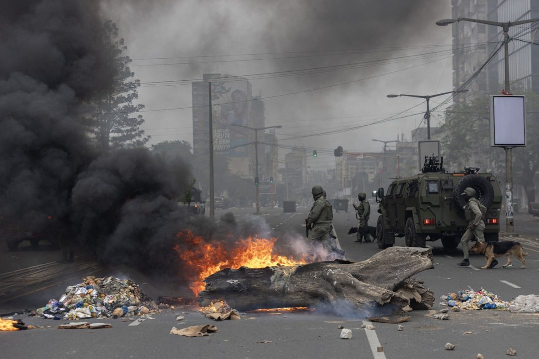 A large log on fire on a city street, alongside piles of litter and trash, with armed soldiers standing in the background next to a dark grey military vehicle