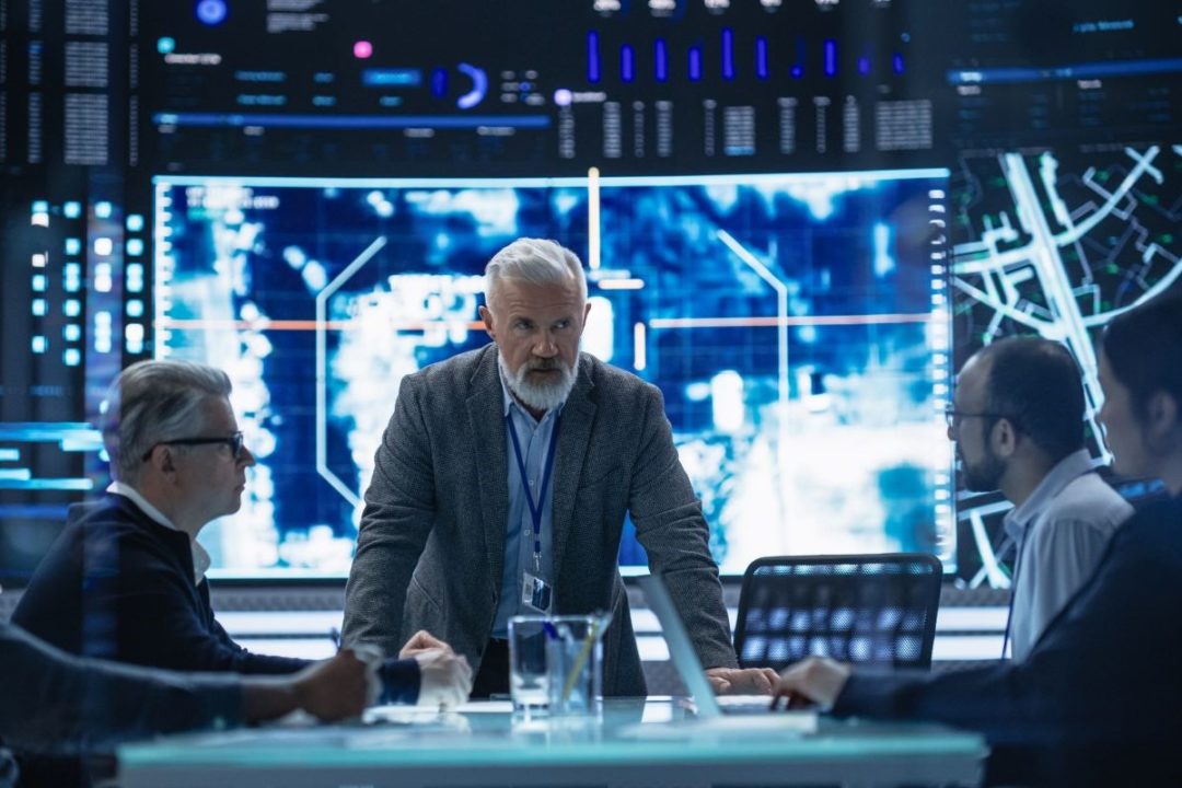 A MAN IN A SUIT STANDS AT THE HEAD OF A TABLE SURROUNDED BY SEATED BUSINESSMEN, A GLOWING BANK OF SCREENS BEHIND HIM