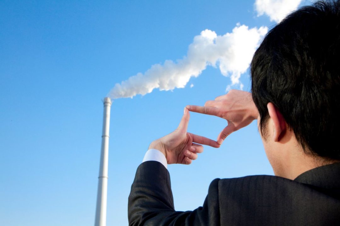 A MAN FORMS HIS THUMBS AND FOREFINGERS INTO A FRAME IN FRONT OF A TALL METAL CHIMNEY BELCHING GASES UNDER A CLEAR BLUE SKY