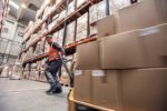 A man in a reflective orange vest and grey pants pulling a cart stacked with brown cardboard boxes, alongside shelves of more boxes stacked to the ceiling of a large warehouse