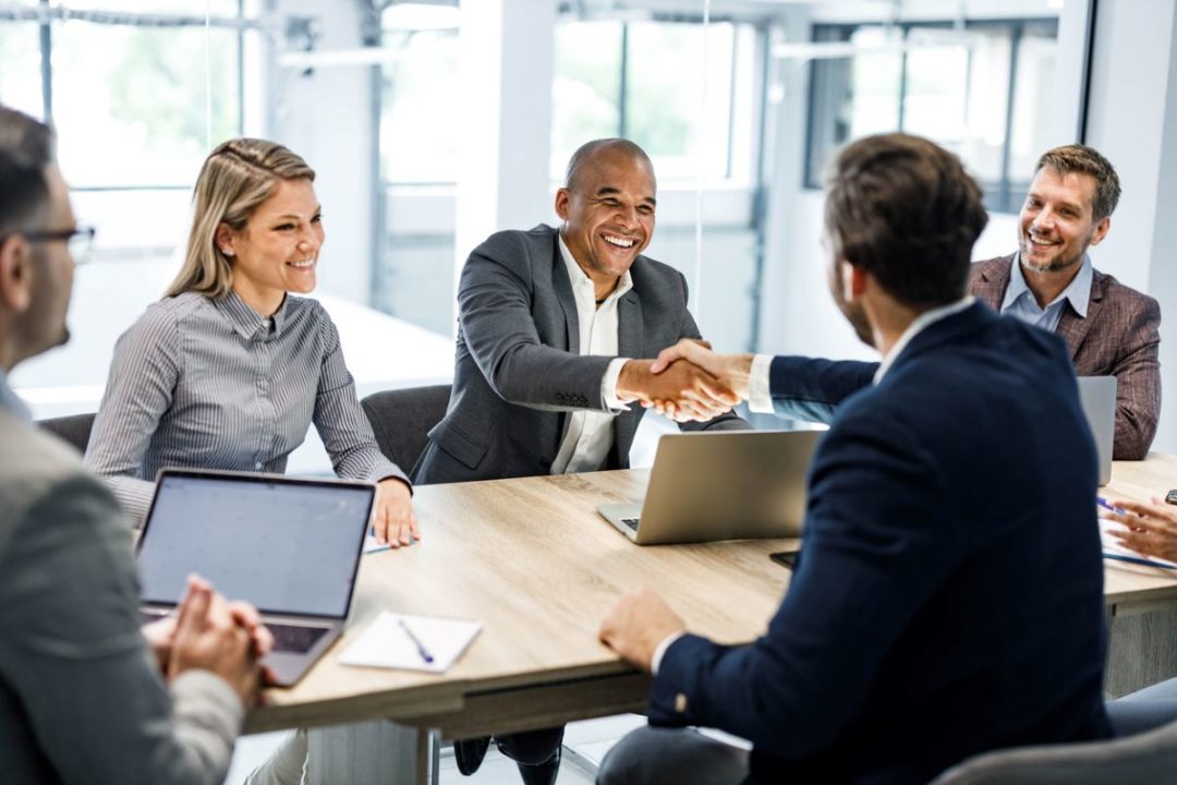 A GROUP OF BUSINESS MEN AND WOMEN SIT AROUND A TABLE, TWO OF THEM SHAKING HANDS, SMILING2.jpg