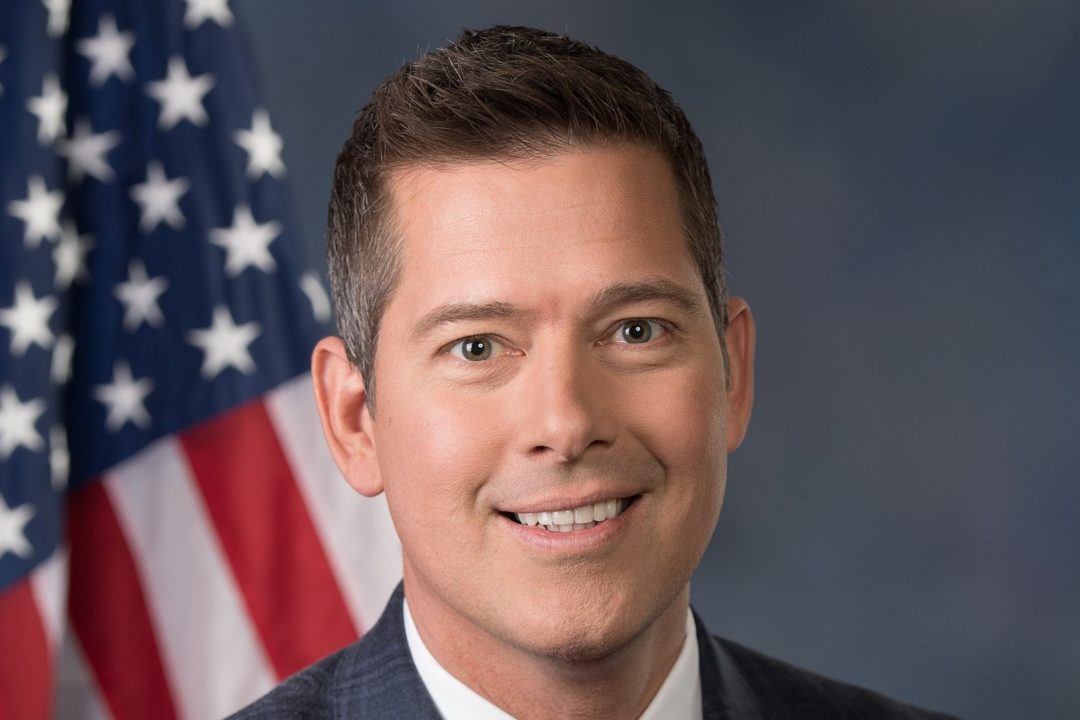 A MIDDLE-AGED WHITE MAN IN A SUIT AND TIE SMILES IN FRONT OF AN AMERICAN FLAG