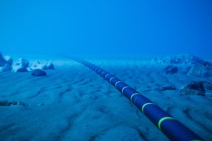 A think blue cable with evenly spaced neon yellow stripes, sitting along a seabed lined with rocky terrain