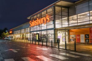 The front of a grocery store with large glass windows, orange walls, and "Sainsbury's" written in large orange neon letters across the top above the entrance.