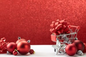 A miniature shopping cart with a large red gift bow on top of it, alongside red orb Christmas ornaments, all set against a glittery red background