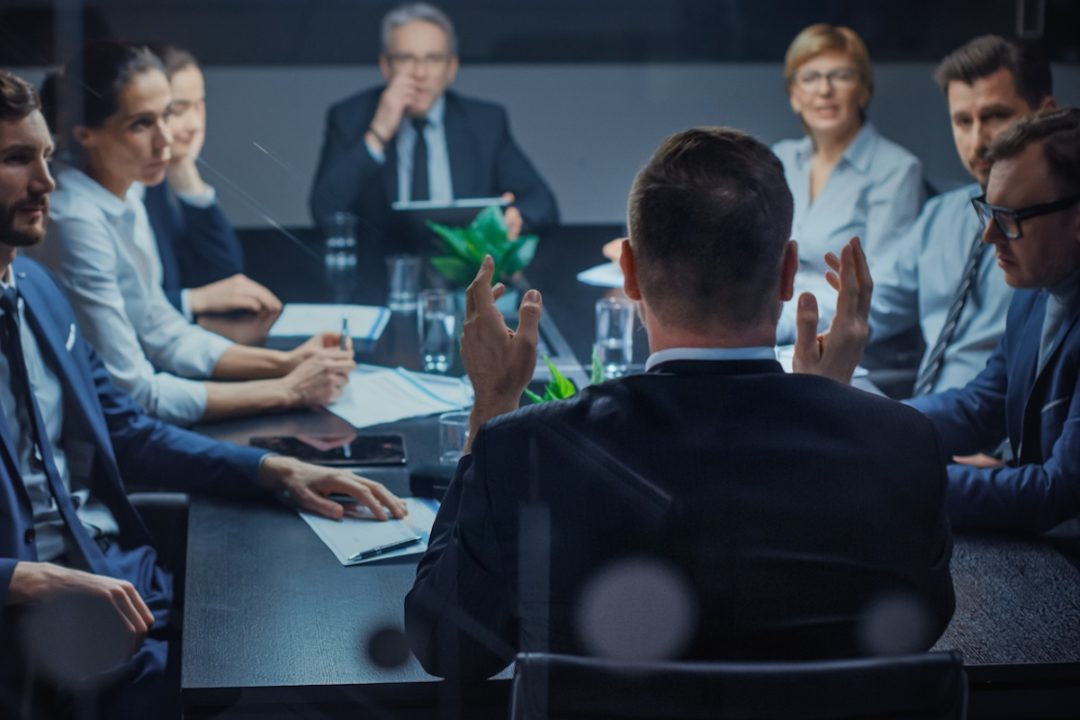 A group of men and women in dress clothes having a discussion while sitting in a dark grey conference room