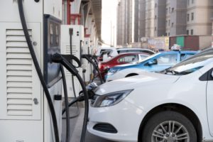 A line of small electric vehicles, each plugged into electric charging stations while parked below a raised freeway.