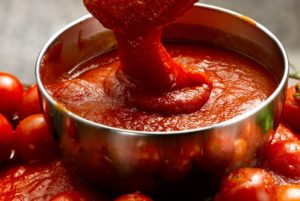 A LADLE POURS THICK TOMATO PUREE INTO A COPPER PAN