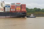 A black container ship seen from the side, with four large stacks of red and grey shipping containers on the front end.