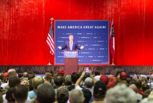 Donald Trump speaking to a crowd while wearing a navy blue suit, set against a blue square background that reads "Make America Great Again," and flanked by American flags on either side.