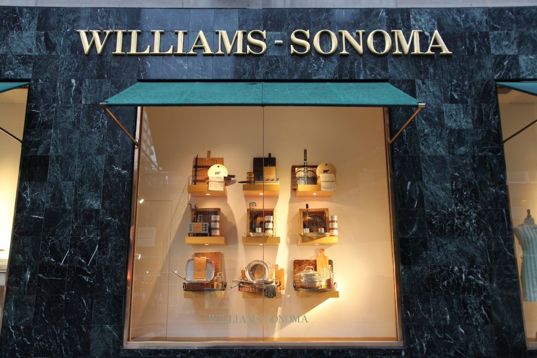 A black marble storefront, with a window in the center with nine square shelves displaying an array of home goods items, with "Williams-Sonoma" written across the top of the storefront in gold embossed lettering.