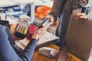 A woman in a grey jacket handing a credit card to a woman wearing a blue sweater, holding a key pad.