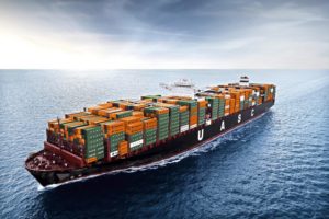 A black container ship at sea carrying stacks of green and orange shipping containers