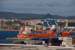 An orange container ship docked below a blue shipping crane and stacks of blue and white shipping containers, with a hillside dotted with red-roofed homes in the background