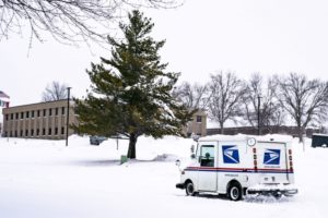 A USPS DELIVERY TRUCK DRIVES UP A SNOWY HILL TOWARDS A LARGE OFFICE BUILDING